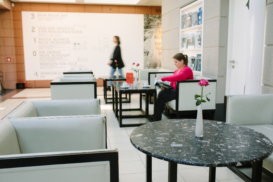 Blick ins Caféfoyer des Deutschen Architekturmuseum mit schwarz-weißen Sessel und Tischen und einer lesenden, sitzenden Dame und einer laufenden Person im Hintergrund