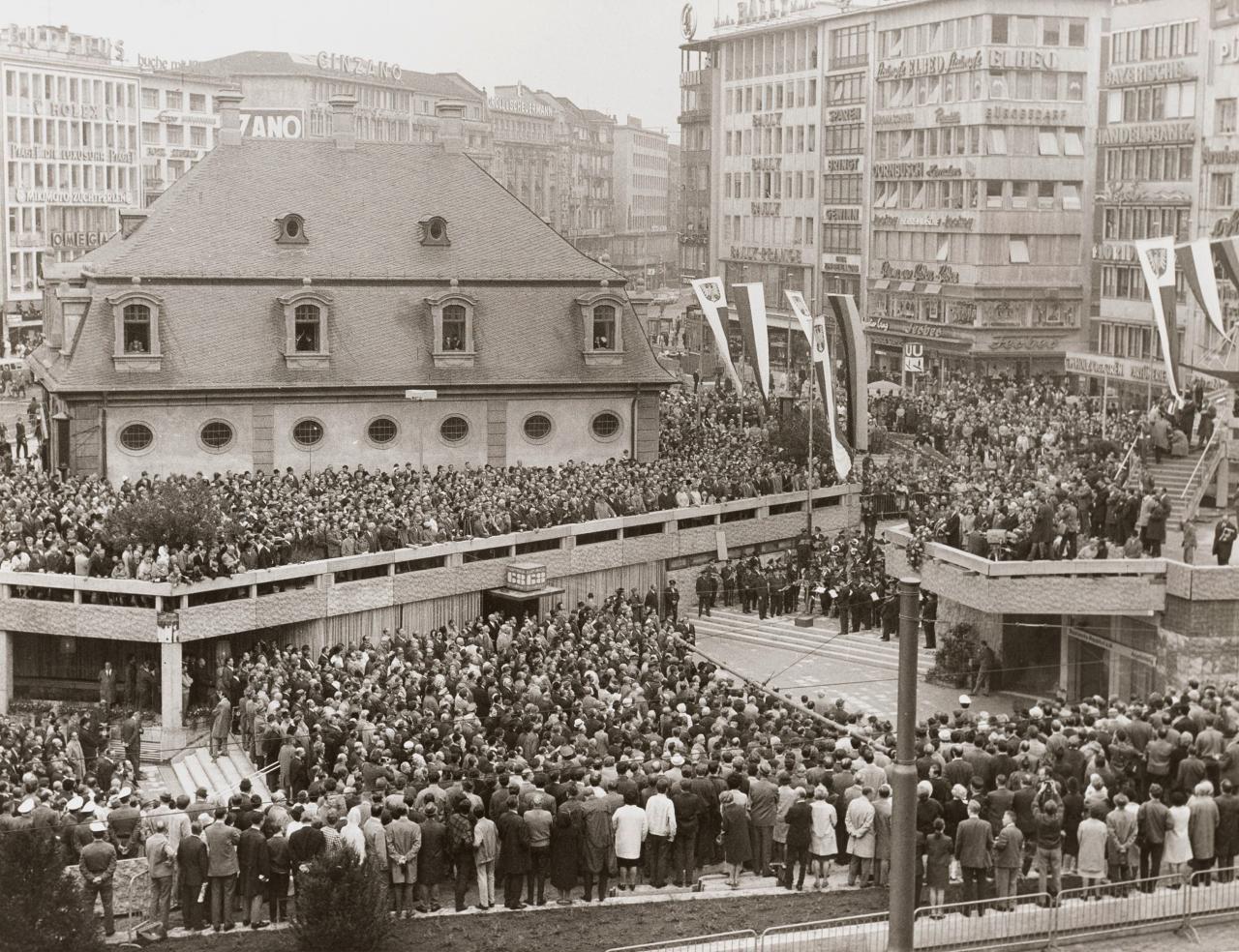 Enlarge image: Schwarz-weiß Fotografie, auf der eine Menschmenge auf einem Platz mit einem Eingang in die B-Ebene vor Häusern steht