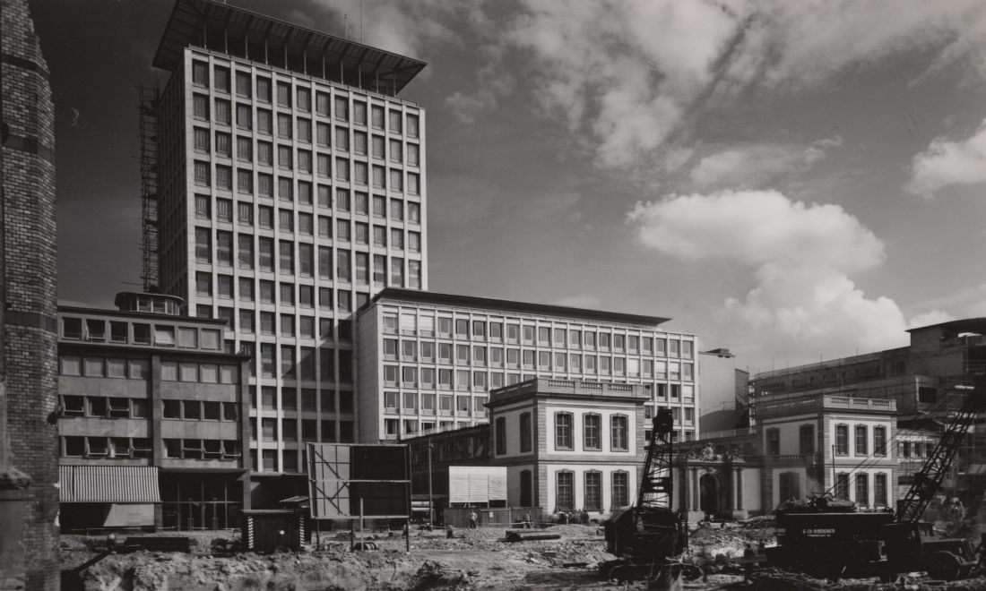 Bild vergrössern: Schwarz-weiß Fotografie mit einem hohen 50-Jahre-Gebäude in der Mitte und weiteren Gebäuden drum herum und einer Baustelle davor