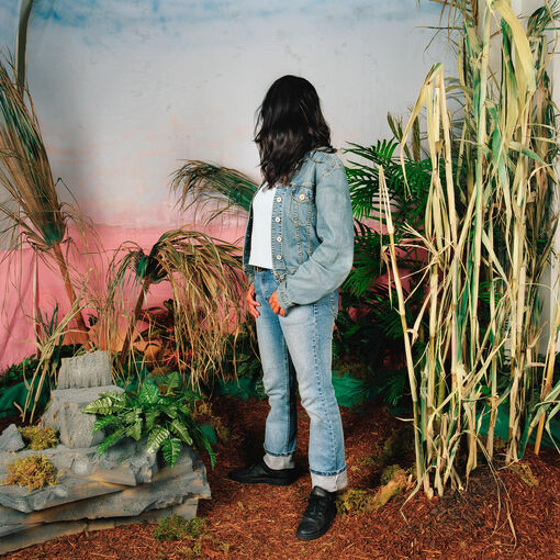 Gina Osterloh, Looking Back I Accepted Your Invitation, 2005, aus der Serie »Somewhere Tropical«. © Gina Osterloh, 2025, courtesy of the artist and Silverlens, Manila and New York Eine Frau in Jeansjacke und -hose steht zwischen Pflanzen und schaut hinter sich auf eine Wand, die wie ein Himmel angestrichen ist