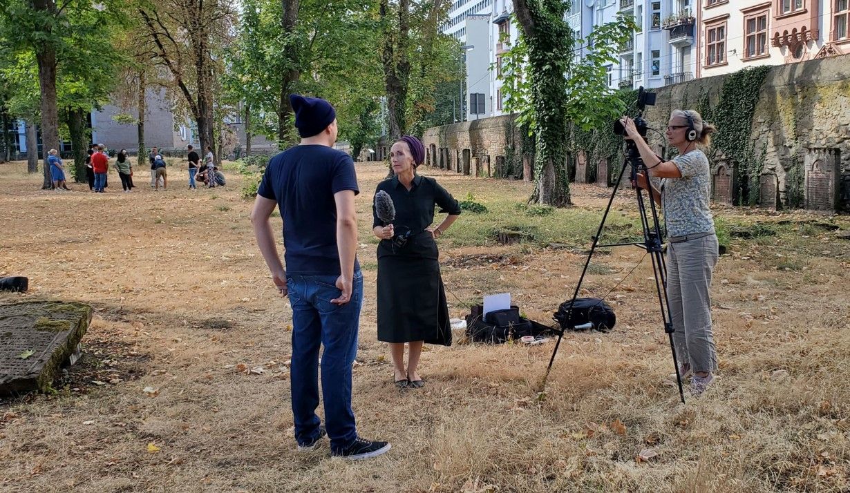 Enlarge image: Foto einer Interviewsituation, ein Mann steht mit dem Rücken zum Betrachter auf einem Friedhof und wird von einer Frau mit Mikro interviewt, eine weitere Frau rechts im Bild filmt