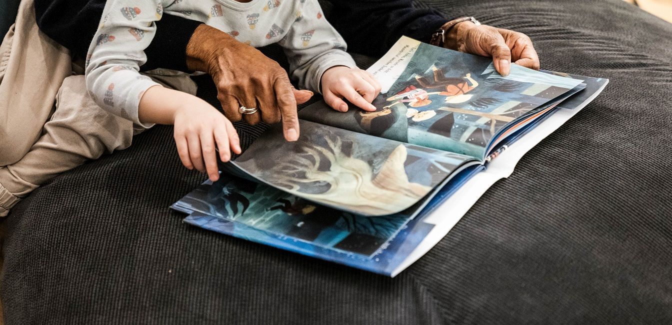 Kinderbuch-Ausstellung 2024 © Klingspor Museum – Offenbach am Main, Foto: Simon Malz Hände von einer älteren Person und einem Kind blättern in einem Buch mit großen Fotos