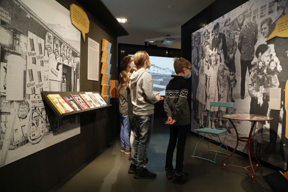 Bild vergrössern: Mehrere Kinder schauen in einem Ausstellungsraum auf ein großes Schwarz-Weiß-Foto an der Wand auf dem Kinder und Männer in schwarzen Uniformen auf der Straße stehen