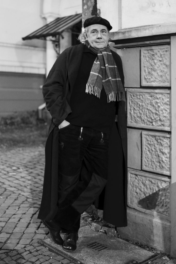 Bild vergrössern: Schwarz-Weiß Foto eines Mannes mit Mantel Schal und Mütze an eine Hauswand gelehnt