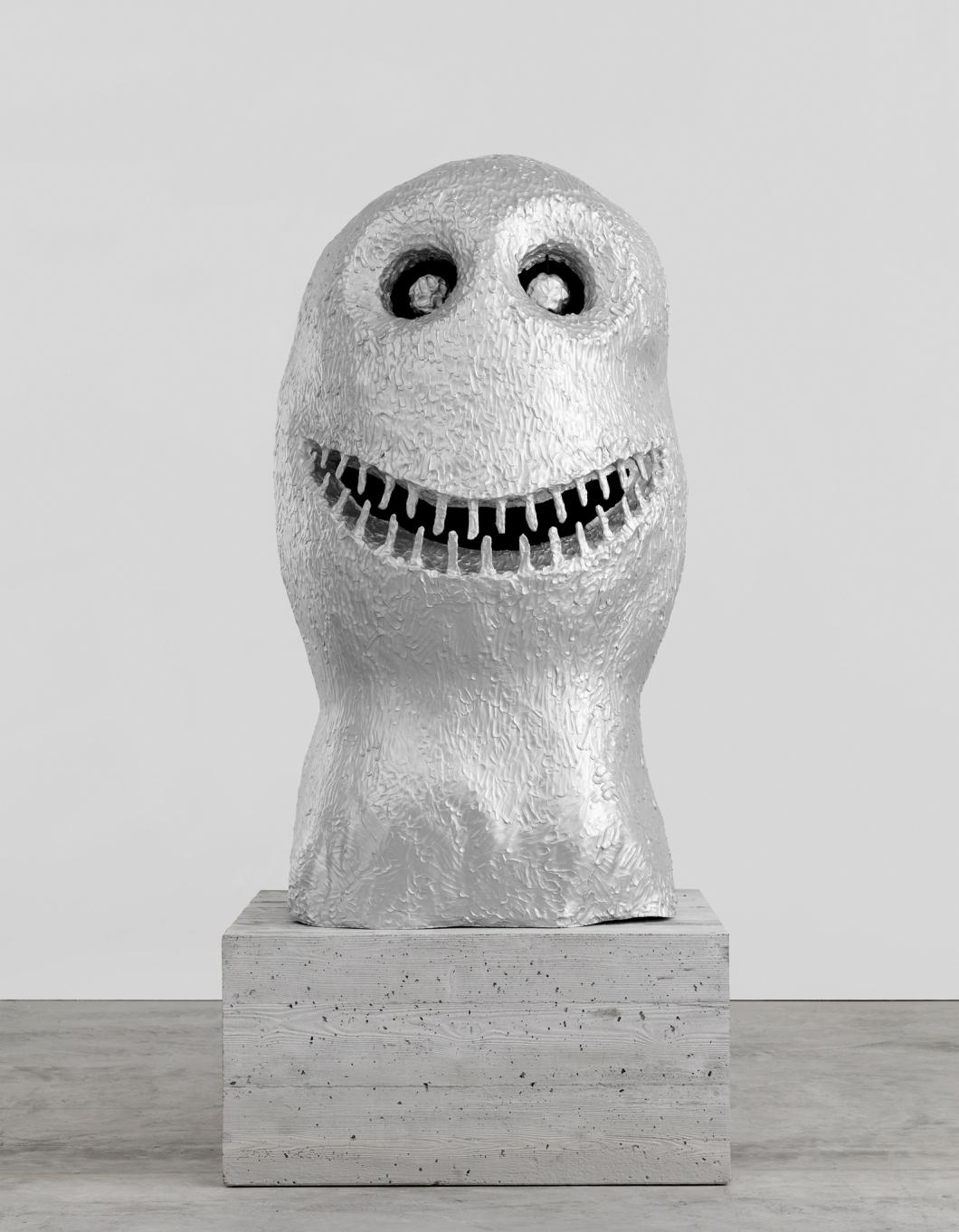Eine Skulptur aus Aluminium, die wie ein vereinfachter grinsender Kopf aussieht, auf einem Betonsockel