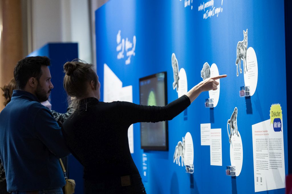 Bild vergrössern: Zwei Personen links im Bild stehen vor einer blauen Wand rechts mit Bildern und Texten, die eine Person zeigt auf etwas