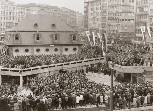 Schwarz-weiß Fotografie, auf der eine Menschmenge auf einem Platz mit einem Eingang in die B-Ebene vor Häusern steht