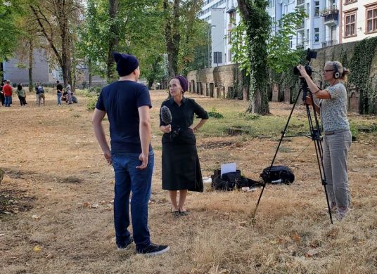 Foto einer Interviewsituation, ein Mann steht mit dem Rücken zum Betrachter auf einem Friedhof und wird von einer Frau mit Mikro interviewt, eine weitere Frau rechts im Bild filmt