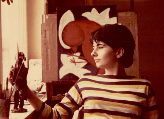 Auf dem sepiafarbenen Foto schaut ein Mädchen auf Pinseln, im Hintergrund ein Gemälde mit einem Vogel