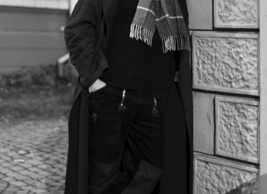 Schwarz-Weiß Foto eines Mannes mit Mantel Schal und Mütze an eine Hauswand gelehnt