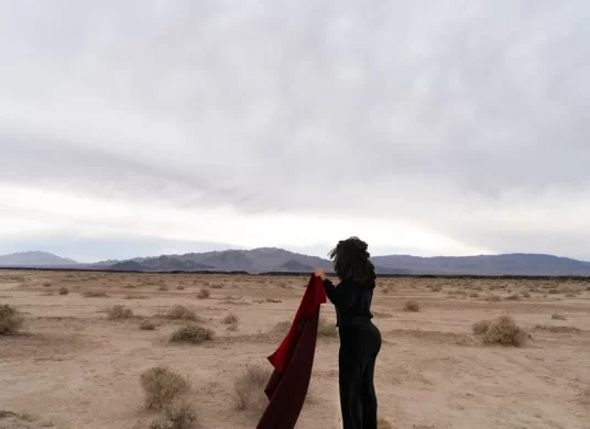 Fotografie, auf der eine schwarzgekleidete Frau in einer kargen Landschaft ein dunkelrotes Tuch in den Händen hält