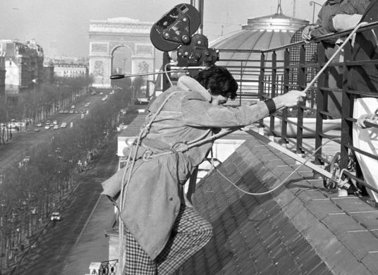 Schwarz-Weiß-Foto eines Kameramannes mit Kamera über der Schulter, der an einem Seil an einem Dach klettert, im Hintergrund der Arc de Triomphe in Paris