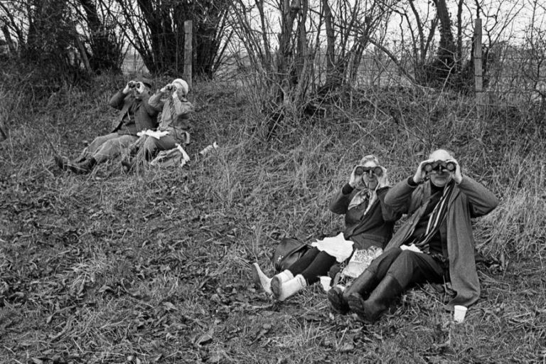 Schwarz-weiß Fotografie, auf der zwei ältere Paare auf einer Wiese unter Gebüsch sitzen und mit Ferngläsern in die Ferne schauen