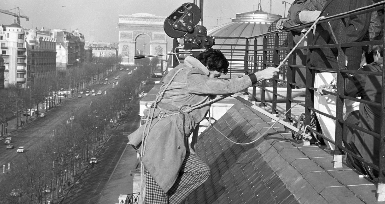 Bild vergrössern: Schwarz-Weiß-Foto eines Kameramannes mit Kamera über der Schulter, der an einem Seil an einem Dach klettert, im Hintergrund der Arc de Triomphe in Paris